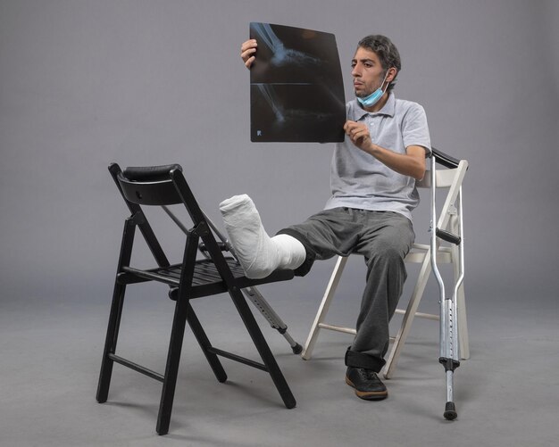 Vista frontal del hombre joven sentado con el pie roto y sosteniendo la radiografía en la pared gris medicina giro dolor masculino pierna rota