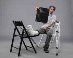 Foto gratuita vista frontal de un hombre joven sentado con un pie roto y sosteniendo una radiografía en el piso gris, dolor de torsión, pierna rota, pie de accidente masculino