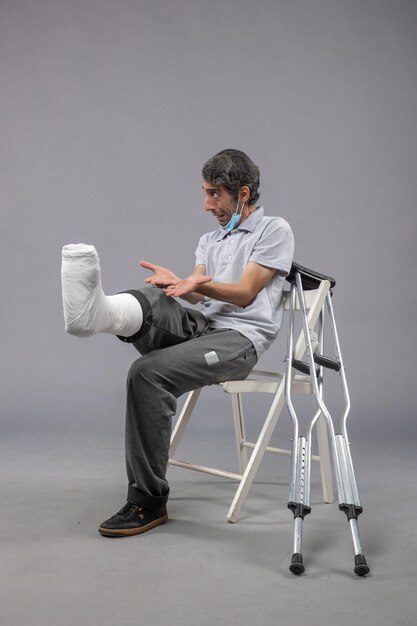 Vista frontal hombre joven sentado con el pie roto vendado en la pared gris accidente de pierna giro dolor en el pie masculino