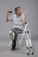 Foto gratuita vista frontal de un hombre joven sentado con un pie roto y un vendaje con una tarjeta bancaria en la pared gris, un dolor en el pie del accidente masculino torcido