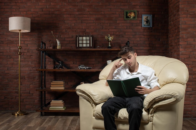 Vista frontal del hombre joven sentado en el sofá leyendo un libro dentro de la habitación trabajo trabajo noche de la casa