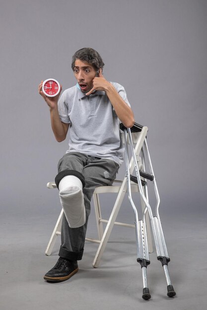 Vista frontal hombre joven sentado con vendaje atado de pie roto y sosteniendo el reloj en la pared gris giro de pie pierna de dolor de accidente masculino