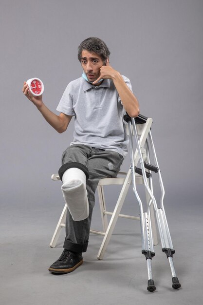Vista frontal hombre joven sentado con vendaje atado de pie roto y sosteniendo el reloj en la pared gris giro de pie pierna de dolor de accidente masculino