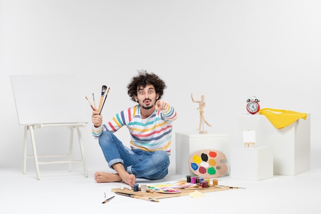 Foto gratuita vista frontal del hombre joven sosteniendo borlas para dibujar en la pared blanca