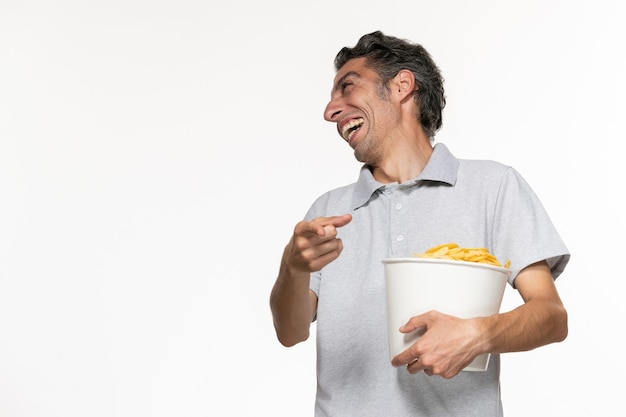 Vista frontal hombre joven sosteniendo una cesta con papas fritas y hablando con alguien riendo sobre una superficie blanca