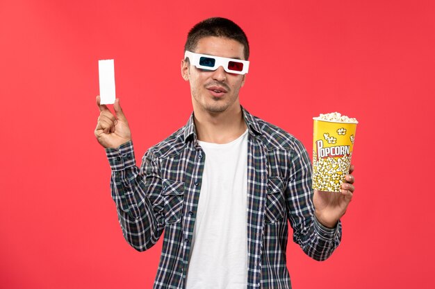 Vista frontal hombre joven sosteniendo palomitas de maíz y billete en -d gafas de sol en la pared roja cine cine película