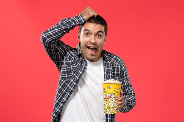 Vista frontal hombre joven sosteniendo el paquete de palomitas de maíz y posando en la pared de color rojo claro cine teatro película película