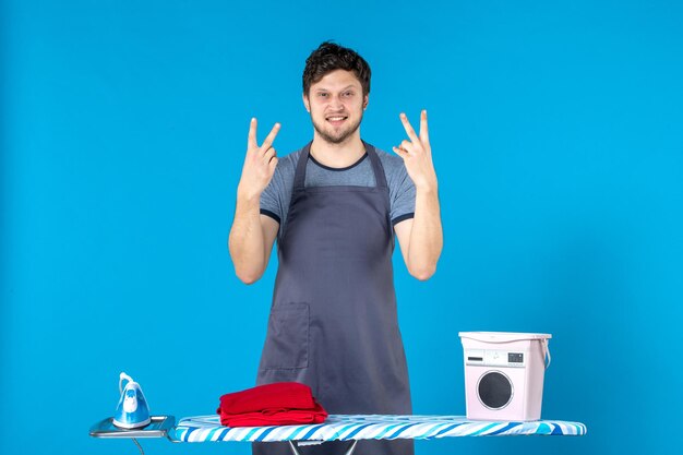 Vista frontal del hombre joven con tabla de planchar sobre fondo azul hombre ropa quehaceres domésticos lavadora limpieza plancha color de lavandería