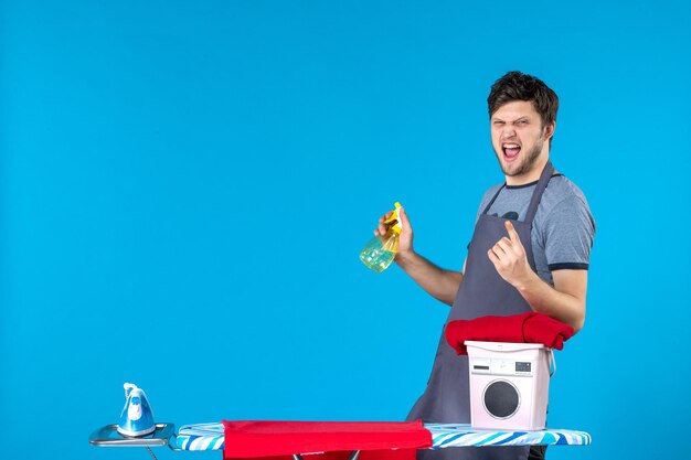 Vista frontal del hombre joven con tabla de planchar sobre fondo azul lavadora color plancha de lavandería quehaceres domésticos hombre