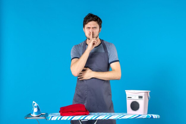 Vista frontal del hombre joven con tabla de planchar sobre fondo azul quehaceres domésticos colores plancha lavadora hombre limpieza