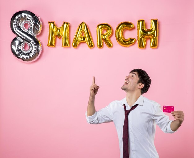 Vista frontal hombre joven con tarjeta bancaria con decoración de marzo sobre fondo rosa color dinero hombre presente igualdad compras vacaciones fiesta femenino