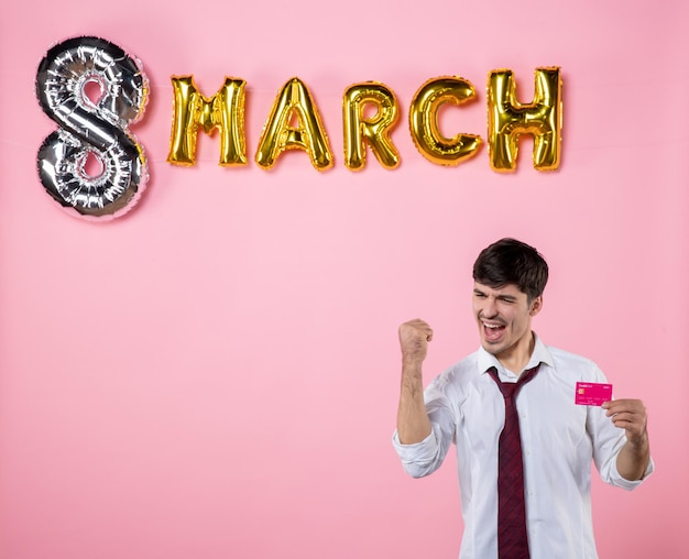 Vista frontal hombre joven con tarjeta bancaria rosa con decoración de marzo sobre fondo rosa dinero hombre presente igualdad compras color fiesta festiva foto femenina