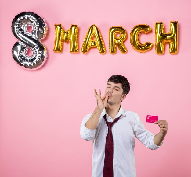 Vista frontal hombre joven con tarjeta bancaria rosa con decoración de marzo sobre fondo rosa fiesta presente vacaciones hombre dinero igualdad color compras