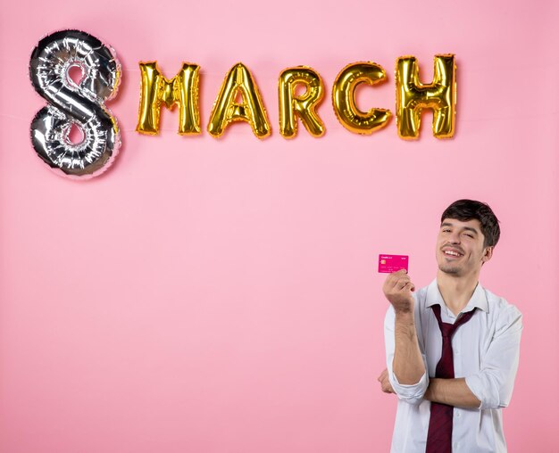 Vista frontal hombre joven con tarjeta bancaria rosa con decoración de marzo sobre fondo rosa igualdad femenina color dinero vacaciones hombre presente compras