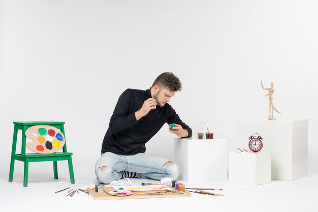 Vista frontal hombre joven trabajando con pinturas en la pared blanca artista pintura arte colores pintura cuadro dibujar foto