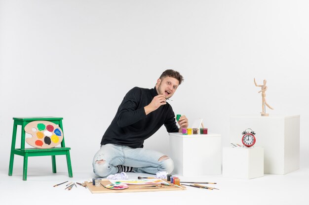 Vista frontal hombre joven trabajando con pinturas en la pared blanca pintura arte color artista pinturas foto imagen dibujar