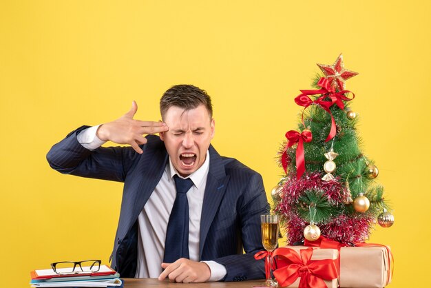 Vista frontal del hombre maravillado manteniendo la pistola de dedos en la sien sentado en la mesa cerca del árbol de Navidad y presenta en amarillo