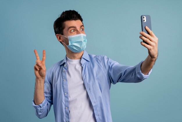 Vista frontal del hombre con máscara médica y tomando selfie mientras hace el signo de la paz