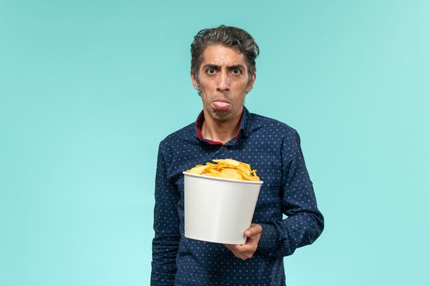 Vista frontal hombre de mediana edad sosteniendo cips y comiendo en superficie azul