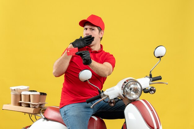 Vista frontal del hombre de mensajería confundido vestido con blusa roja y guantes de sombrero en máscara médica entregando orden sentado en scooter mostrando tarjeta bancaria