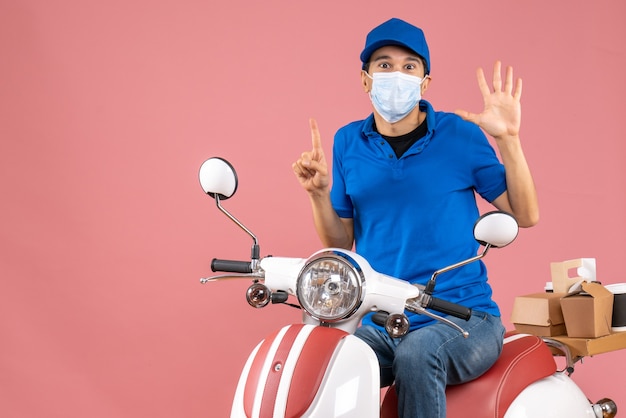 Vista frontal del hombre de mensajería en máscara médica con sombrero sentado en scooter mostrando cinco apuntando hacia arriba sobre fondo melocotón pastel