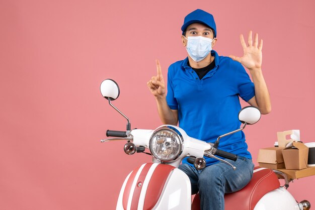 Vista frontal del hombre de mensajería en máscara médica con sombrero sentado en scooter mostrando cinco apuntando hacia arriba sobre fondo melocotón pastel