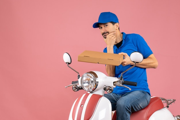 Vista frontal del hombre de mensajería de pensamiento con sombrero sentado en scooter manteniendo orden sobre fondo de melocotón pastel