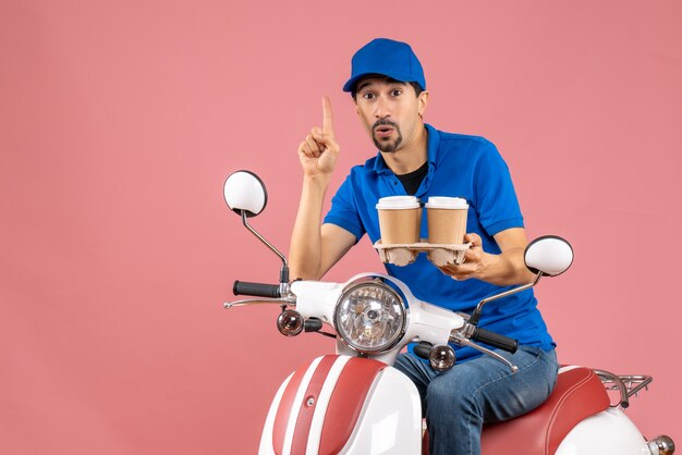 Vista frontal del hombre de mensajería con sombrero sentado en scooter apuntando hacia arriba sobre fondo melocotón pastel