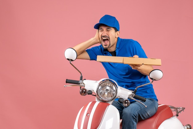 Vista frontal del hombre de mensajería con sombrero sentado en scooter que sufre de dolor de oído sobre fondo de melocotón pastel