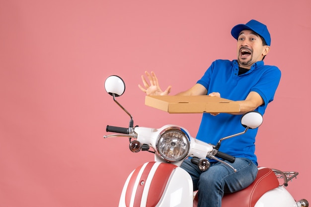 Vista frontal del hombre de mensajería con sombrero sentado en scooter sintiéndose ansioso sobre fondo melocotón pastel