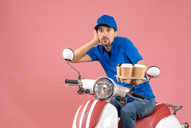 Vista frontal del hombre de mensajería con sombrero sentado en scooter sintiéndose conmocionado sobre fondo melocotón pastel