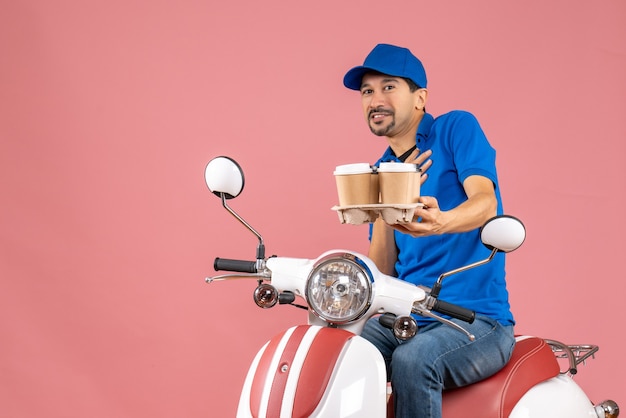 Vista frontal del hombre de mensajería con sombrero sentado en scooter sintiéndose orgulloso sobre fondo melocotón pastel