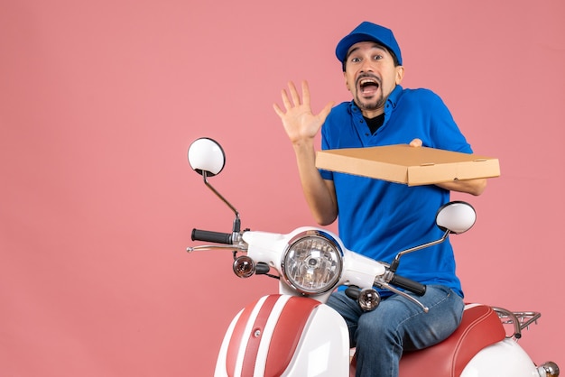 Vista frontal del hombre de mensajería con sombrero sentado en scooter sintiéndose sorprendido sobre fondo melocotón pastel