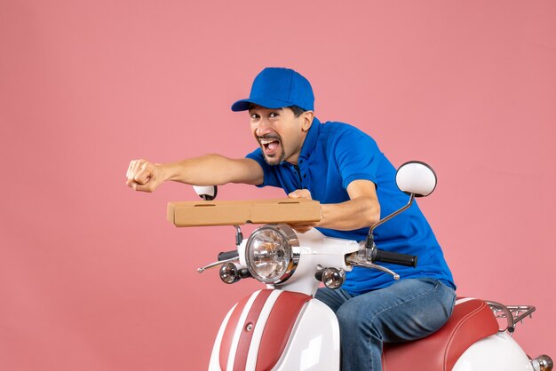 Vista frontal del hombre de mensajería sonriente feliz con sombrero sentado en scooter con orden sobre fondo de melocotón pastel