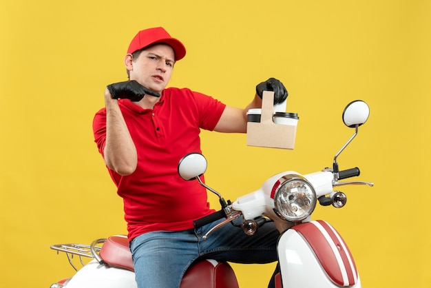 Vista frontal del hombre de mensajería sorprendido vestido con blusa roja y guantes de sombrero en máscara médica entregando orden sentado en scooter señalando órdenes