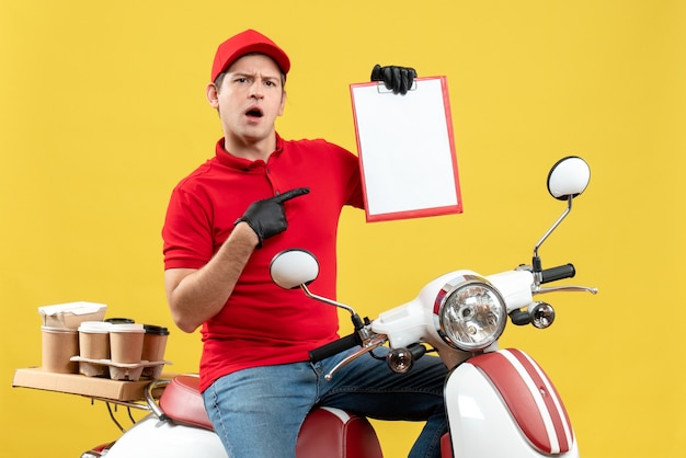 Foto gratuita vista frontal del hombre de mensajería sorprendido vestido con blusa roja y guantes de sombrero en máscara médica entregando orden sentado en scooter sosteniendo documento