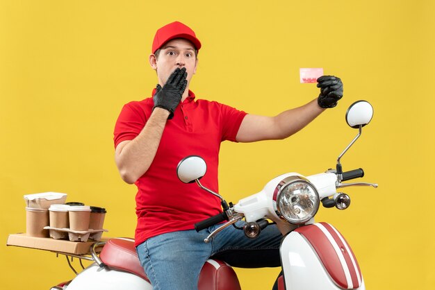 Vista frontal del hombre de mensajería sorprendido vestido con blusa roja y guantes de sombrero en máscara médica entregando orden sentado en scooter con tarjeta bancaria