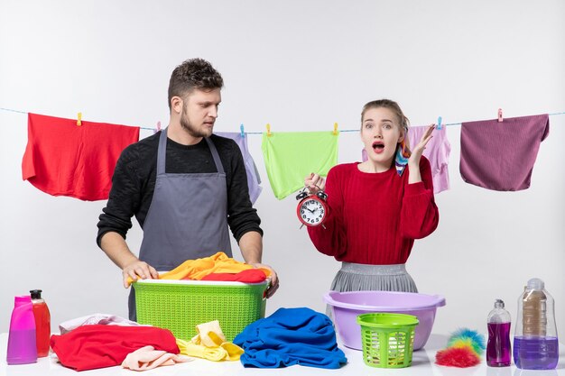 Vista frontal del hombre mirando el despertador y su esposa de pie detrás de la mesa cestas de ropa y lavando cosas en la mesa