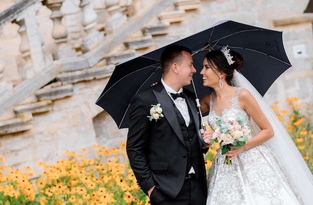 Vista frontal de un hombre y una mujer casados sonriendo y mirándose mientras están de pie bajo un paraguas bajo la lluvia en el fondo de un lecho de flores y escaleras de un edificio antiguo