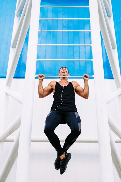 Vista frontal del hombre musculoso guapo sonriente haciendo pull ups, vistiendo ropa deportiva negra.