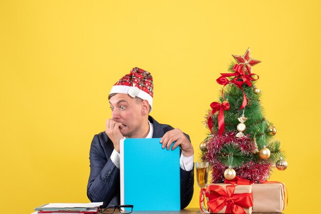 Vista frontal del hombre de negocios agitado sentado en la mesa cerca del árbol de Navidad y presenta en la pared amarilla