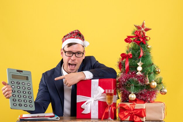 Vista frontal del hombre de negocios apuntando a la calculadora sentado en la mesa cerca del árbol de Navidad y presenta en amarillo