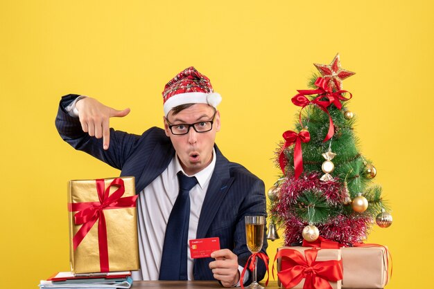 Vista frontal del hombre de negocios apuntando a su regalo sentado en la mesa cerca del árbol de Navidad y presenta en amarillo