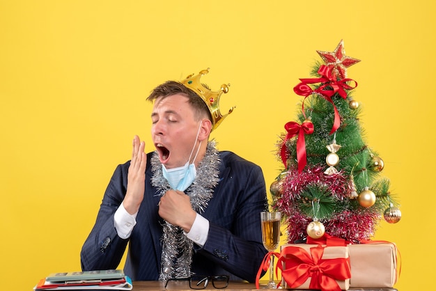 Vista frontal del hombre de negocios bostezando sentado en la mesa cerca del árbol de Navidad y presenta en amarillo