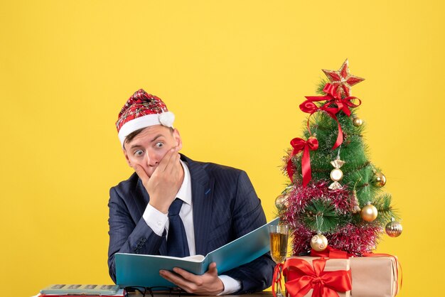 Vista frontal del hombre de negocios confundido sentado en la mesa cerca del árbol de Navidad y presenta en amarillo