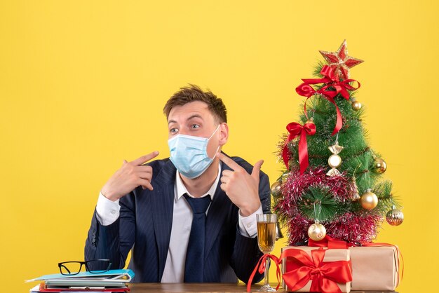 Vista frontal del hombre de negocios confundido sentado en la mesa cerca del árbol de Navidad y presenta en amarillo