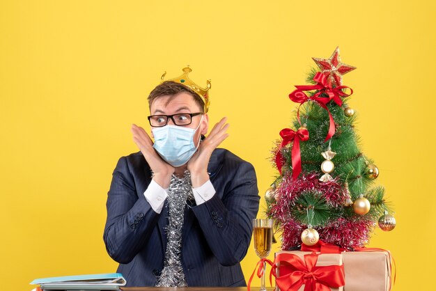 Vista frontal del hombre de negocios con corona sentado en la mesa cerca del árbol de Navidad y presenta en amarillo