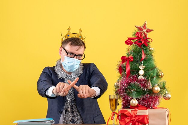 Vista frontal del hombre de negocios cruzando los dedos sentado en la mesa cerca del árbol de Navidad y presenta en la pared amarilla