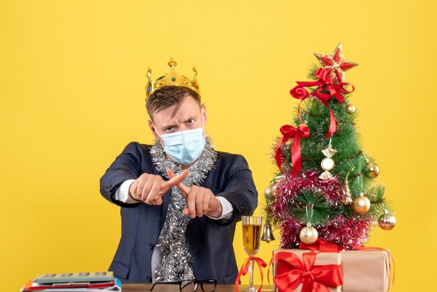 Vista frontal del hombre de negocios cruzando los dedos sentado en la mesa cerca del árbol de Navidad y presenta en la pared amarilla