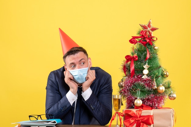 Vista frontal del hombre de negocios deprimido con gorra de fiesta sentado en la mesa cerca del árbol de Navidad y presenta en amarillo